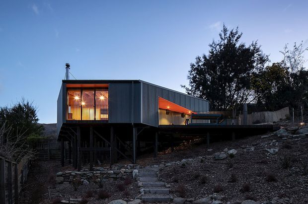 Exterior dusk view of a contemporary elevated house on a steep, rocky site. The dark corrugated-clad structure is raised on slender supports, with warm light glowing through large glazed openings and beneath a covered outdoor terrace. A linear roof form, exposed supporting structure, timber screening and stone retaining walls are visible.