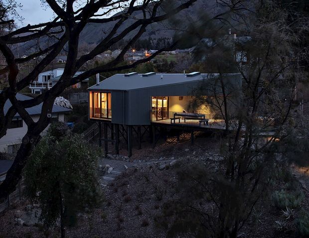 A contemporary elevated house viewed from outside at dusk, set on a steep, wooded hillside. The compact dark metal-clad building has a low-pitched roof, extensive warm-lit glazing, and a timber deck projecting over the rocky terrain. Nearby homes and mountainous bushland are visible in the background.