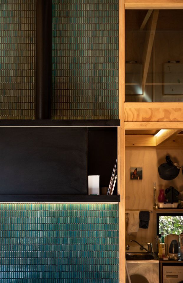 Close-up interior view of a compact, highly detailed built-in joinery installation. Deep teal-green narrow vertical tiles cover the wall and continue below a black-framed recessed display and media niche. Warm honey-toned timber shelving and cabinetry are visible to the right, opening onto a small kitchenette or utility area with a sink and appliances. The composition features strong material contrast between dark cabinetry, colourful tilework and warm timber, with integrated warm lighting.