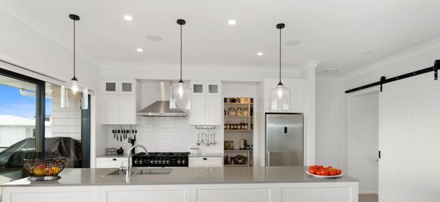 Bright, spacious white kitchen with a large island, white shaker-style cabinetry, pale stone benchtops, stainless-steel appliances, pendant lights, and a black-framed sliding barn-style door. A dining or living area is visible through the glazing at left.