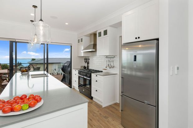 Bright white kitchen with shaker-style cabinetry, a large pale benchtop island, stainless-steel refrigerator and range, pendant lighting, and light timber flooring. Full-height glazing opens to an outdoor terrace with a coastal view.