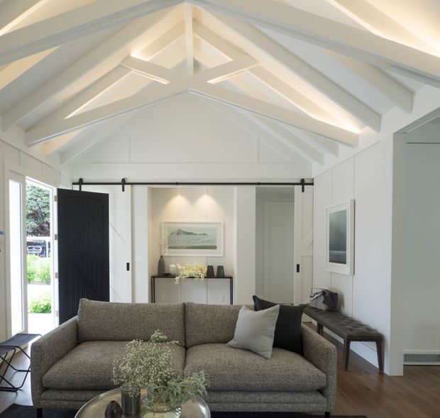 Bright, refined living room with a large textured sectional sofa, dark accent cushions, a slim console table, artwork, and a black sliding barn-style door. The room features white walls, warm timber flooring, and a prominent vaulted ceiling with exposed white-painted rafters and beams, creating an airy contemporary interior.