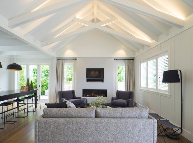 Bright open-plan living room with a dramatic white vaulted ceiling and exposed-style painted beams. The space includes a large grey sectional sofa, two dark lounge chairs, a wall-mounted television above a linear fireplace, a dining area with black pendant lighting, and extensive windows with white plantation shutters. Timber flooring and soft neutral furnishings create a clean, contemporary interior.