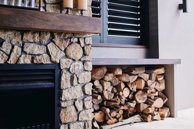 Close-up of a rustic stone fireplace and integrated firewood storage niche. Irregular natural stone surrounds a dark fireplace opening, with a substantial timber mantel above. A horizontal stone or concrete shelf caps the neatly stacked firewood, while dark louvred window shutters and pale rendered walls appear in the background.