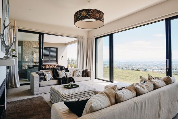 Bright, expansive living room with floor-to-ceiling corner glazing overlooking a broad landscape and city view. The room features two large cream upholstered sofas, a checked ottoman, patterned cushions, pale curtains, dark timber flooring, and a large floral drum pendant light. A stone-clad wall and adjoining outdoor or circulation area are visible beyond the glazing.