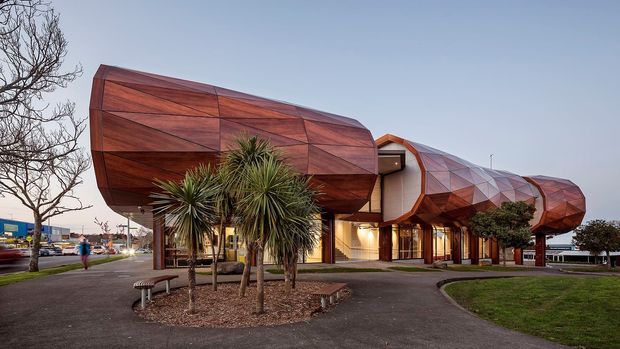 Exterior view of a distinctive contemporary commercial building with an elongated, sculptural form clad in faceted reddish-brown panels. The angular, folded facade and projecting volumes create a striking geometric composition, with glazed entrances and warm interior lighting beneath. Paved paths, landscaped planting, and mature trees surround the building.