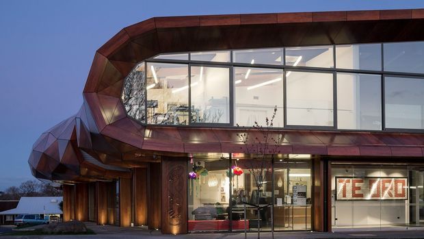 Exterior view of a distinctive contemporary commercial building at dusk, featuring angular faceted weathered-metal cladding, a projecting upper-level glazed volume, extensive black-framed windows, and a recessed illuminated retail or hospitality entrance. Warm interior lighting and timber-toned metal surfaces contrast with the cool evening sky.