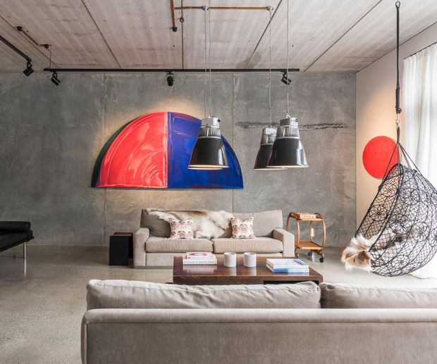 A stylish industrial-contemporary living room with polished concrete flooring, raw grey concrete walls and ceiling, exposed surface-mounted lighting, and suspended black pendant lamps. Two pale upholstered sofas face a dark wood coffee table and a large bold red-and-blue abstract wall artwork. A sculptural hanging lounge chair sits beside sheer-curtained glazing, with warm timber accents and a small black grand piano at the edge of the room.