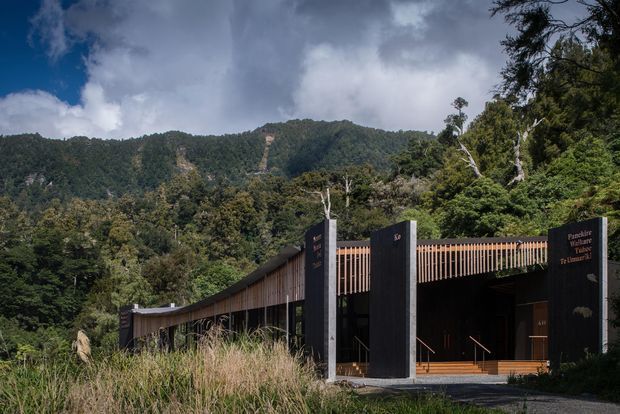 A low, elongated contemporary building set within dense native forest, with steep mountain ridges rising behind it. The dark facade is framed by tall charcoal vertical piers and warm timber screening, creating a sheltered entrance and covered walkway. The architecture is carefully integrated into the heavily vegetated, mountainous landscape under a dramatic cloudy sky.
