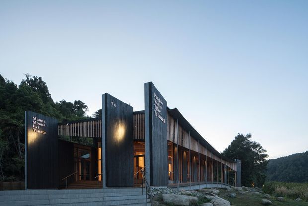 Exterior architectural photograph of a long, low-slung building set into a wooded, sloping landscape. The facade combines dark vertical solid panels with extensive warm timber screening, tall glazed openings, and a gently curved or sloping roofline. Broad concrete steps and exposed natural rock lead to the entrance, while warm interior lighting glows through the glazing at dusk.