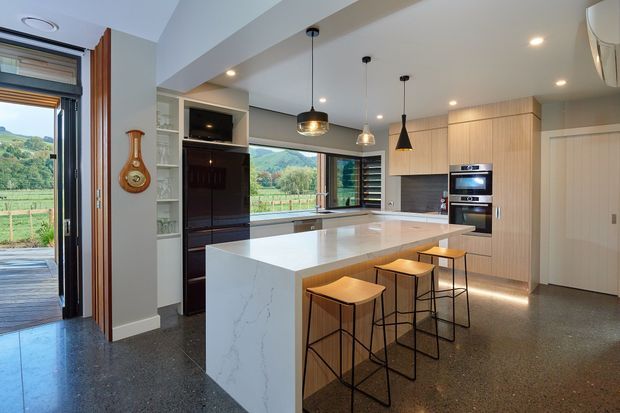 Contemporary open-plan kitchen with a large white stone island and integrated seating, dark timber cabinetry, pale timber wall cabinetry, built-in appliances, pendant lighting, and polished concrete or terrazzo flooring. The kitchen opens toward a rural landscape through large windows and a glazed doorway.