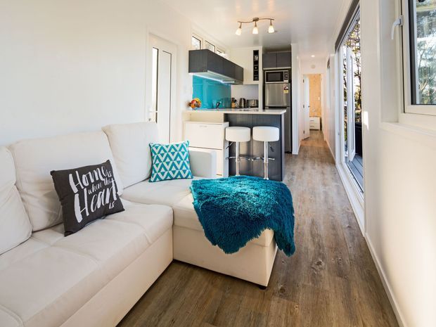 Bright, compact open-plan interior combining a living room and galley kitchen. A large white sectional sofa occupies the foreground, with a turquoise throw and patterned cushion. The kitchen has white cabinetry, dark upper cabinets and benchtops, a vivid turquoise splashback, stainless-steel appliances, and two bar stools. Timber-look flooring runs through the space toward a narrow hallway, with abundant natural light from large windows and glazed doors.