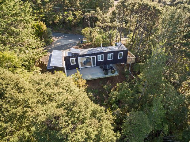 Aerial view of a compact dark-clad house nestled among dense trees on a sloped, wooded site. The building has a long rectangular form, a corrugated metal pitched roof, several white-framed windows, and a broad elevated outdoor deck or terrace facing the surrounding vegetation. A driveway and utility lines are visible behind the house.