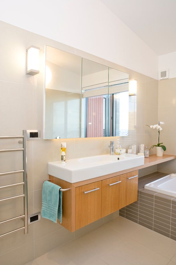 Bright contemporary bathroom with a wall-mounted timber vanity, integrated white basin, large mirrored cabinet, beige tiled walls and floor, and a tiled bathtub surround. A stainless-steel heated towel rail, wall sconces, chrome tapware, and a small orchid complete the clean, functional space. The mirror reflects a window and exterior view.