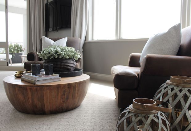 A bright, softly styled living room with a large round wood coffee table, upholstered armchairs, layered neutral textiles, floor-to-ceiling curtains, and a dark media wall. Decorative woven lanterns and a central planter add texture and warmth.