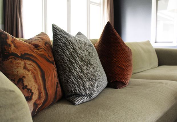Close-up of a softly upholstered light beige sofa with several decorative cushions in burnt orange marbled fabric, charcoal herringbone weave, and deep rust textured fabric. Tall windows with dark curtains provide bright natural light in the background.