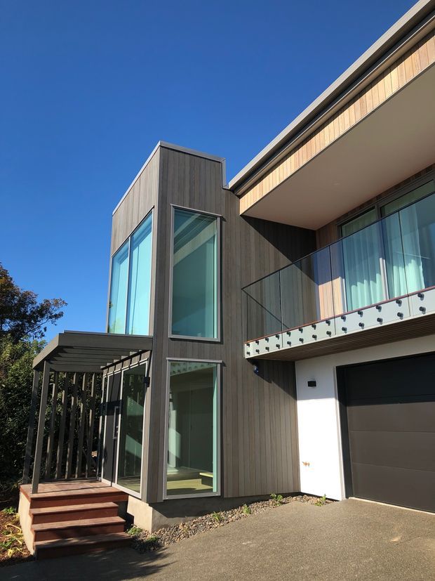 A contemporary multi-level residential facade with dark vertical cladding, warm timber-toned upper cladding, expansive blue-tinted glazing, a glass-railed balcony, and an integrated dark garage door. A covered entry porch with timber steps sits beside the projecting glazed volume, viewed from the driveway under clear daylight.
