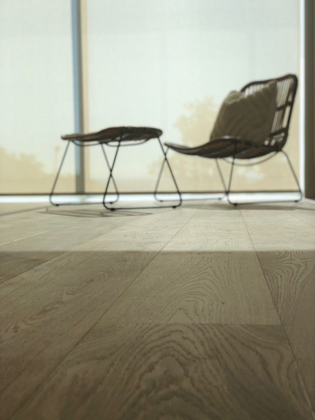 A softly lit, minimalist interior with pale wide-plank timber flooring, a black woven lounge chair and matching ottoman positioned in front of large translucent windows or blinds. The low camera angle emphasizes the natural wood grain and airy, uncluttered atmosphere.