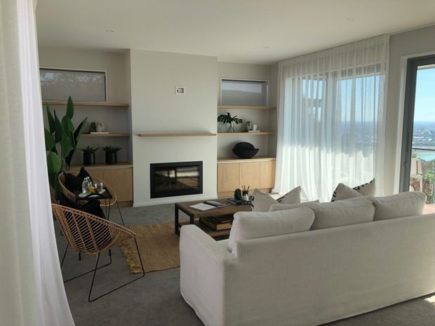 Bright contemporary living room with a large white sectional sofa facing a built-in fireplace. Full-height sheer curtains frame expansive glazing with a coastal or elevated view beyond. Built-in timber shelving and cabinetry flank the fireplace, while a woven lounge chair, low timber coffee table, neutral rug, and indoor plants add warmth and texture.