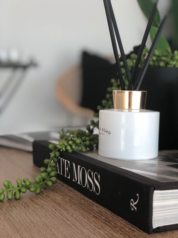 Close-up styled interior vignette on a wood-grain tabletop, featuring a white cylindrical reed diffuser with a metallic brass-gold cap, a trailing green plant, and stacked black design books. The background is softly blurred and appears to be a contemporary living or display setting.
