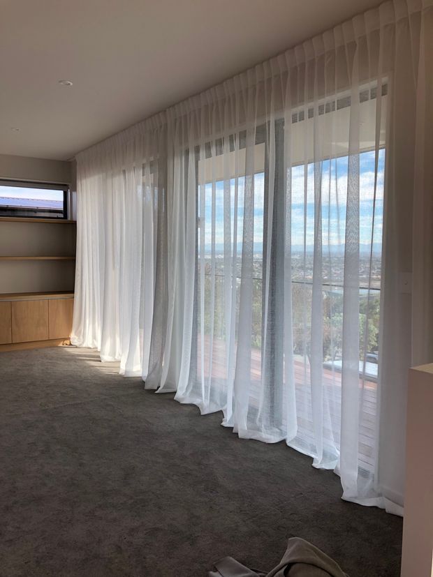 A bright, mostly empty interior room with floor-to-ceiling sheer white curtains spanning a wall of glazing and opening to a balcony or terrace with an elevated city view. The room has grey carpet, a warm timber built-in shelving and cabinetry unit, recessed ceiling lights, and a calm contemporary feel.