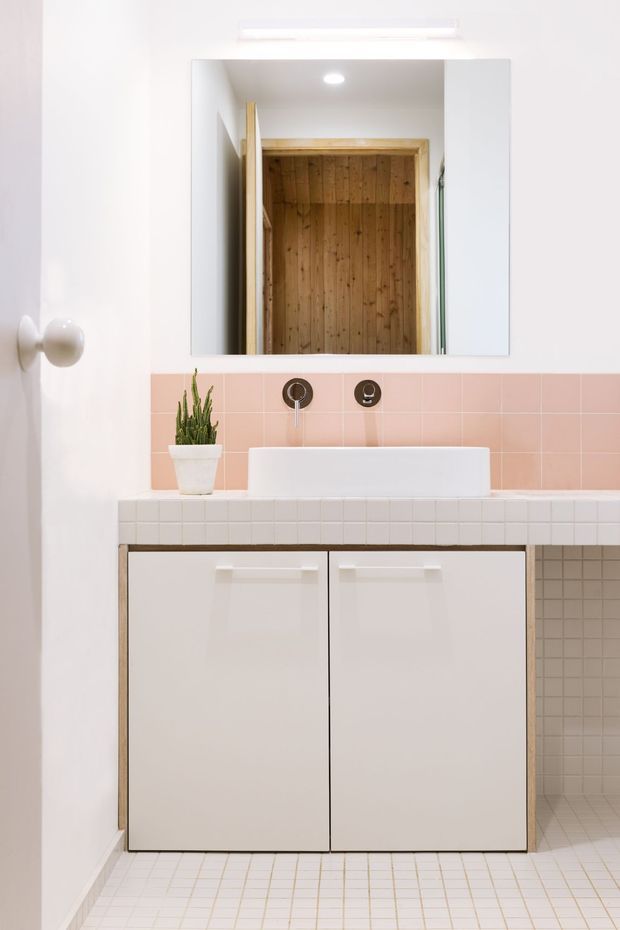 A bright, compact bathroom vanity with a white vessel basin, wall-mounted dark metal tapware, pale pink square tiled splashback, white tiled benchtop and floor, and simple white cabinetry. A large rectangular mirror reflects a warm timber-lined doorway or adjacent space. A small potted plant adds greenery.