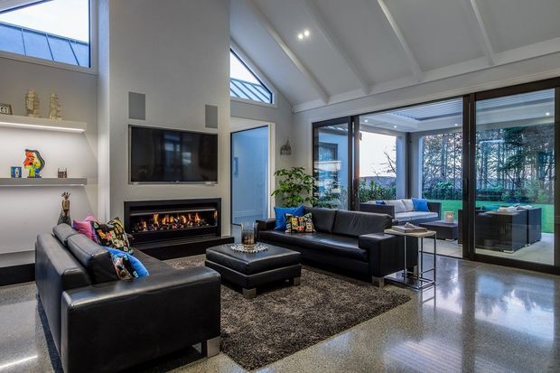 Contemporary living room with black leather sofas, a large rug and ottoman, built-in gas fireplace and television wall, extensive glazing, and a high pitched coffered ceiling. The polished concrete floor and indoor-outdoor connection create a spacious, refined entertaining area.