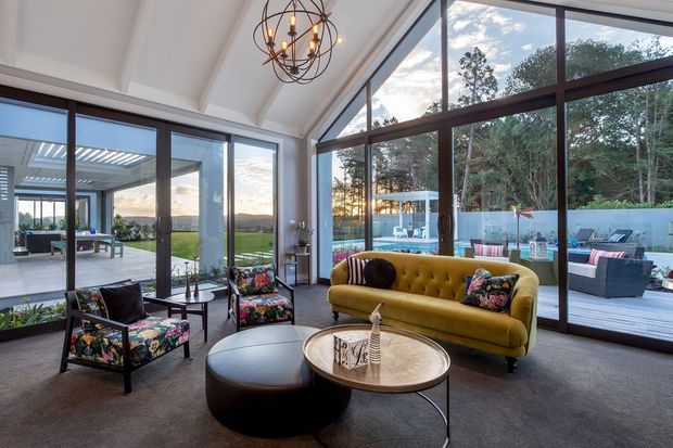 A bright, spacious living room with a dramatic vaulted ceiling and full-height dark-framed glazing on multiple sides. The room features a mustard tufted sofa, patterned lounge chairs, round coffee tables, carpeted flooring, and a sculptural spherical pendant light. Outdoor entertaining areas, a pool, and landscaped surroundings are visible through the glazing.
