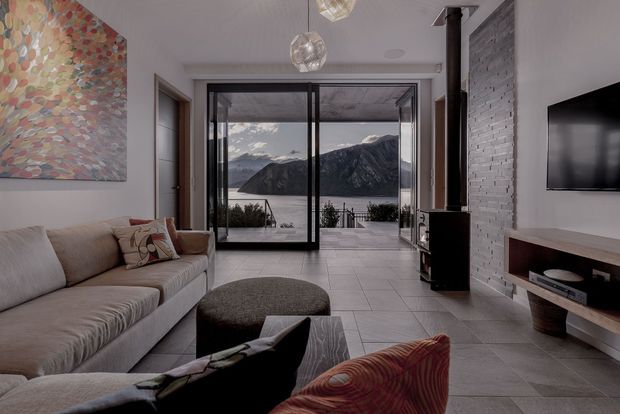A contemporary living room with a large sectional sofa, round ottoman, wood coffee table, wall-mounted television and suspended glass pendant lights. The room has large black-framed sliding glass doors opening to a terrace with a dramatic mountain and water view. Interior finishes include large pale floor tiles, a white-painted brick feature wall, and a dark freestanding wood stove with a vertical flue.