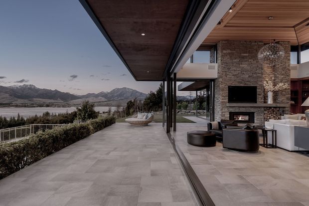 A contemporary open-plan living room opening onto a broad terrace through full-height sliding glazing. The space features a large stacked-stone fireplace wall with a recessed television, warm timber soffit and ceiling lining, pale large-format stone or porcelain flooring, dark slim window frames, and modern neutral seating. The terrace overlooks a lake and mountain landscape.