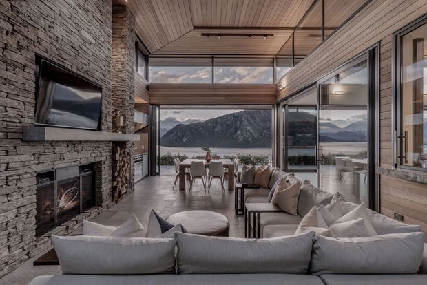A spacious contemporary living room with a large sectional sofa arranged around a stone-clad fireplace and wall-mounted television. Floor-to-ceiling glazing and clerestory windows frame a dramatic mountain and lakeside view, while warm timber-lined walls and ceiling, dark-framed openings, and a dining area create a refined indoor-outdoor connection.