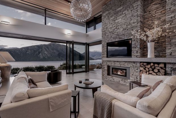 A sophisticated living room with expansive floor-to-ceiling glazing overlooking a lake and rugged mountains. The room features a dramatic full-height stacked-stone fireplace wall with integrated television and timber log storage, a linear timber-slat ceiling, pale upholstered sofas and armchairs, dark occasional tables, and a sculptural spherical pendant light. The open glazing and restrained neutral palette create a refined, warm contemporary retreat.