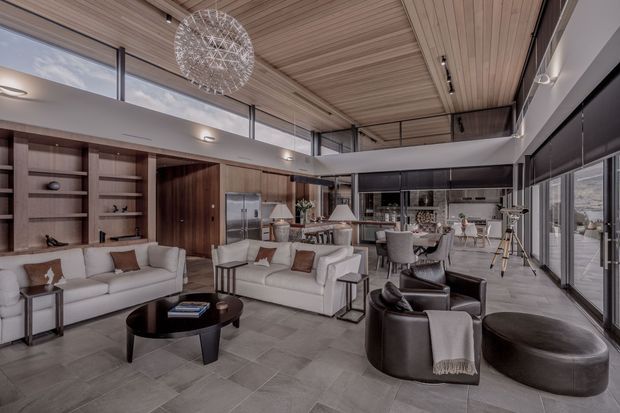 A spacious, refined open-plan living area with a double-height ceiling and extensive floor-to-ceiling glazing. The room combines white sofas, dark leather lounge chairs, a round black coffee table, built-in timber shelving and cabinetry, a dining area, and a kitchen in the background. A linear timber ceiling, large spherical pendant light, and pale stone-look floor create a warm contemporary interior with strong indoor-outdoor connections.