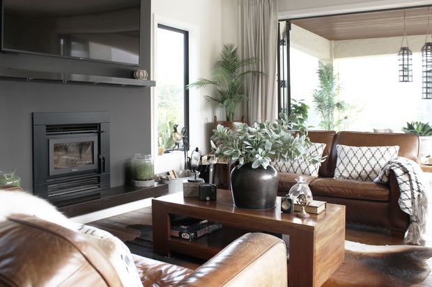 A bright, warmly styled living room with a dark built-in fireplace and television wall, leather sofas and armchairs, a timber coffee table, patterned cushions, indoor plants, and large glazed openings leading to an outdoor area. The space has a relaxed, refined lodge-inspired feel with strong contrast between dark joinery and light walls.