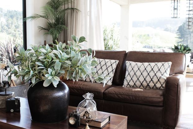 Bright, airy living room with a brown leather sofa, patterned cushions, abundant indoor plants, and a large black ceramic planter arranged around a timber coffee table. Floor-to-ceiling glazing brings in natural light and views of greenery, while pendant lights and neutral curtains complete the relaxed, eclectic interior.