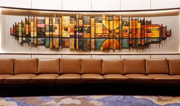 A refined lounge or hospitality seating area featuring a long brown leather banquette beneath a large colourful abstract geometric wall artwork. Warm recessed lighting highlights the artwork and upholstered seating, while a patterned carpet adds muted blue, beige and mauve tones.