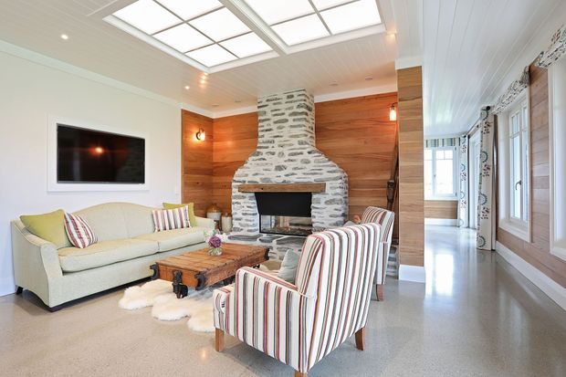 Bright, inviting living room with a large white-and-grey stone fireplace as the focal point, warm timber wall panelling, a wall-mounted television, upholstered sofa and striped armchairs arranged around a rustic wooden coffee table. The room features polished concrete flooring, a white paneled ceiling with large skylights, and tall windows with patterned curtains along one side.