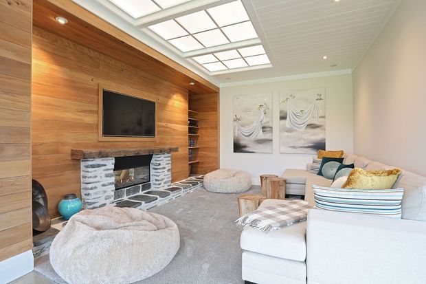 Bright, cozy living room with a large white sectional sofa, oversized floor cushions, and a timber feature wall incorporating a wall-mounted television, open shelving, and a rustic stone-faced fireplace. A large skylight or glazed ceiling panel brings in natural light, while neutral furnishings, soft textiles, and coastal-toned artwork create a relaxed interior.