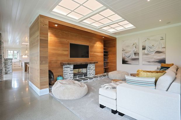 Bright contemporary living room with a large built-in timber feature wall, recessed television, open shelving, and a stone-clad fireplace. A white sectional sofa, soft ottoman, pale rug, and layered cushions furnish the room. A large illuminated ceiling panel provides a skylight-like focal point, while a hallway and kitchen area are visible beyond.