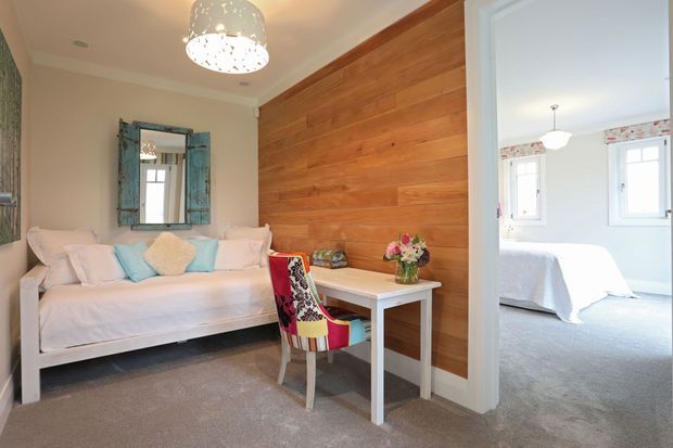 A bright, compact sitting room with a white sofa, pastel cushions, a small white desk, and a colourful upholstered chair. A full-height warm timber feature wall provides contrast against pale walls and carpet, while a distressed blue mirror and patterned pendant light add decorative character. Through the open doorway, a bright bedroom with white furnishings is visible.