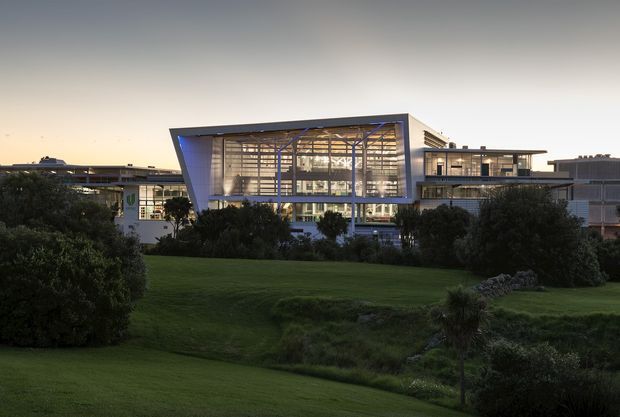 Exterior dusk view of a large contemporary institutional or commercial building set within landscaped grounds. The architecture features a prominent glazed central atrium with visible structural framing, angular projecting roof or facade elements, and connected low-rise wings with flat roofs. Dark greenery and lawns occupy the foreground.