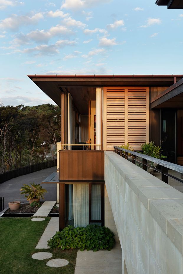 A contemporary multi-level residential building viewed from an elevated outdoor walkway. The facade combines warm timber cladding and horizontal timber screening with dark-framed glazing, broad projecting roof eaves, and a pale masonry retaining wall. Balconies and railings overlook a landscaped sloping site with trees, lawn, stepping stones, and low planting. The image has a warm, refined architectural character under a bright blue sky.