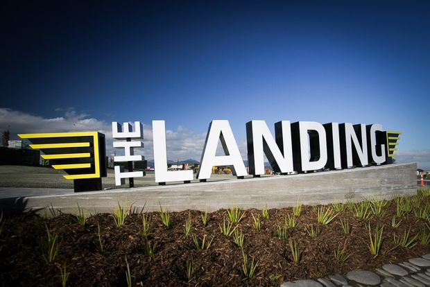Exterior landmark signage reading “The Landing,” with large white three-dimensional letters and bold black-and-yellow graphic elements mounted on a concrete plinth. The sign sits within a newly planted landscaped bed under a bright blue sky.