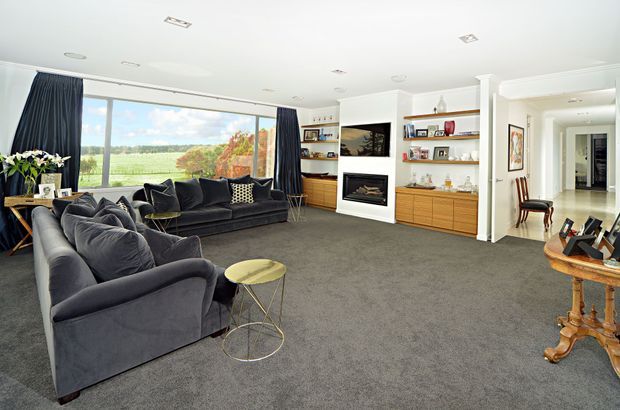 Spacious, bright living room with dark charcoal upholstered sofas arranged around a small round brass-toned side table. A large window provides views of open countryside, while built-in timber shelving and cabinetry frame a fireplace and display objects. The room also includes dark curtains, grey carpet, white walls and ceiling, recessed lighting, and a hallway opening to the right.
