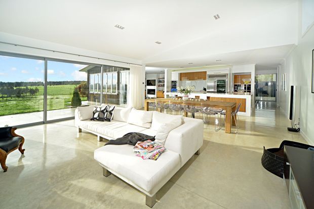 Bright, expansive open-plan living area with floor-to-ceiling glazing overlooking a green rural landscape. A white sectional sofa defines the living zone, while a timber dining table and chairs sit between the lounge and a contemporary kitchen with a central island. Polished pale flooring, white walls, recessed lighting, and abundant daylight create an airy, refined interior.