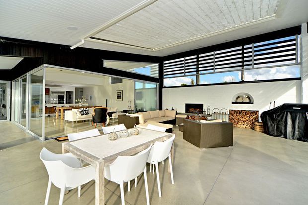 Bright open-plan contemporary living and dining area with polished concrete flooring, extensive glazing, black horizontal louvres, a white-lined ceiling, and a striking indoor fireplace with stacked timber storage. A large dining table and upholstered seating occupy the foreground, while a kitchen and living area are visible beyond the glazed partition.