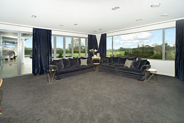 Spacious contemporary living room with floor-to-ceiling windows overlooking a green landscape. The room features dark charcoal sofas, matching floor-length curtains, patterned cushions, small side tables, recessed ceiling lights, and wall-to-wall grey carpet.