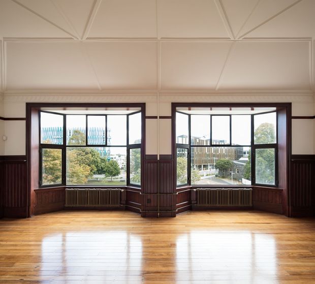 An empty, bright heritage-style interior with two large angled bay windows overlooking an urban setting. The room features polished timber flooring, dark stained timber wall panelling and window surrounds, ornate painted ceiling panels with geometric detailing, and original cast-iron radiators beneath the windows.