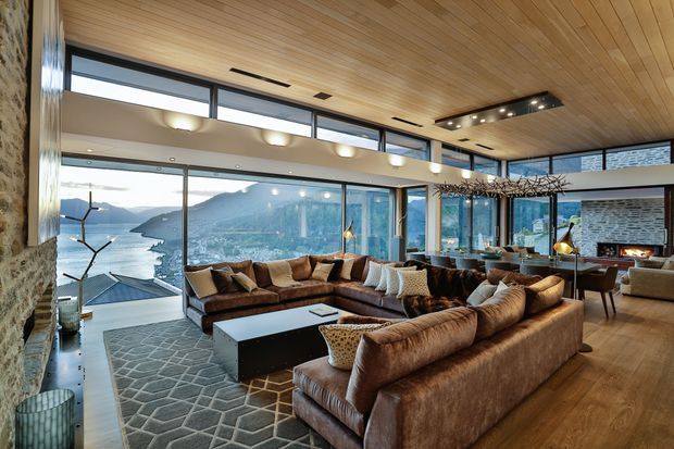 A spacious contemporary living room with floor-to-ceiling glazing overlooking a mountainous waterfront landscape. The room features a warm timber-lined ceiling and timber flooring, a large brown leather sectional seating arrangement, a dark rectangular coffee table, a patterned area rug, exposed stone walls, built-in lighting, and a dining area with a stone fireplace visible at the rear.