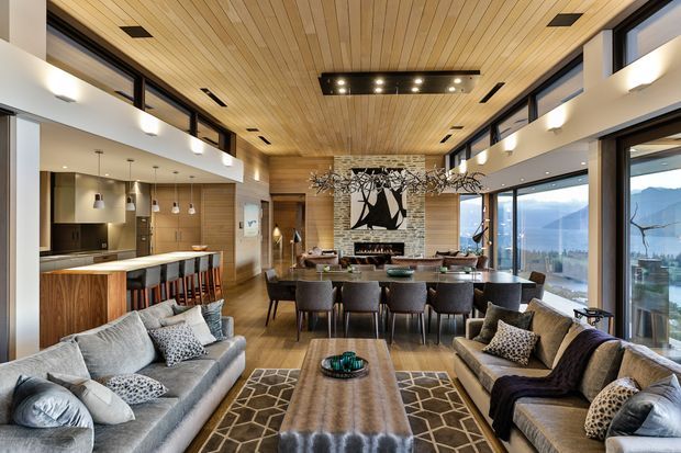 Spacious open-plan living and dining area with a contemporary kitchen, featuring a warm timber-lined ceiling, extensive glazing, stone and timber finishes, two large sofas, a dining table, and a kitchen island with bar seating. Mountain scenery is visible through the windows.